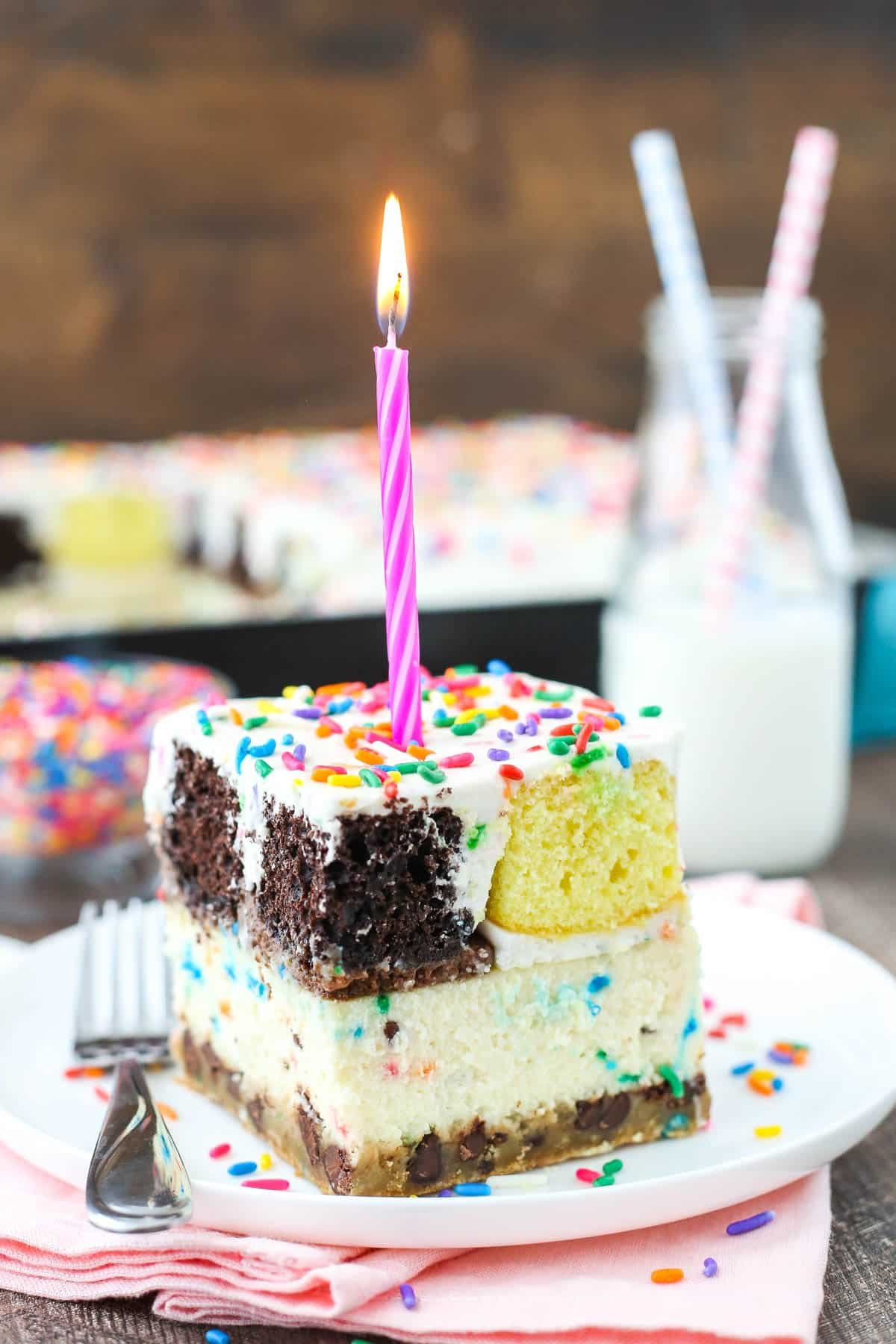 This epic birthday cake combines chewy chocolate chip cookies, creamy Funfetti cheesecake, moist cupcakes & fluffy cake batter whipped cream! slice of ultimate birthday cake on white plate with pink candle on top