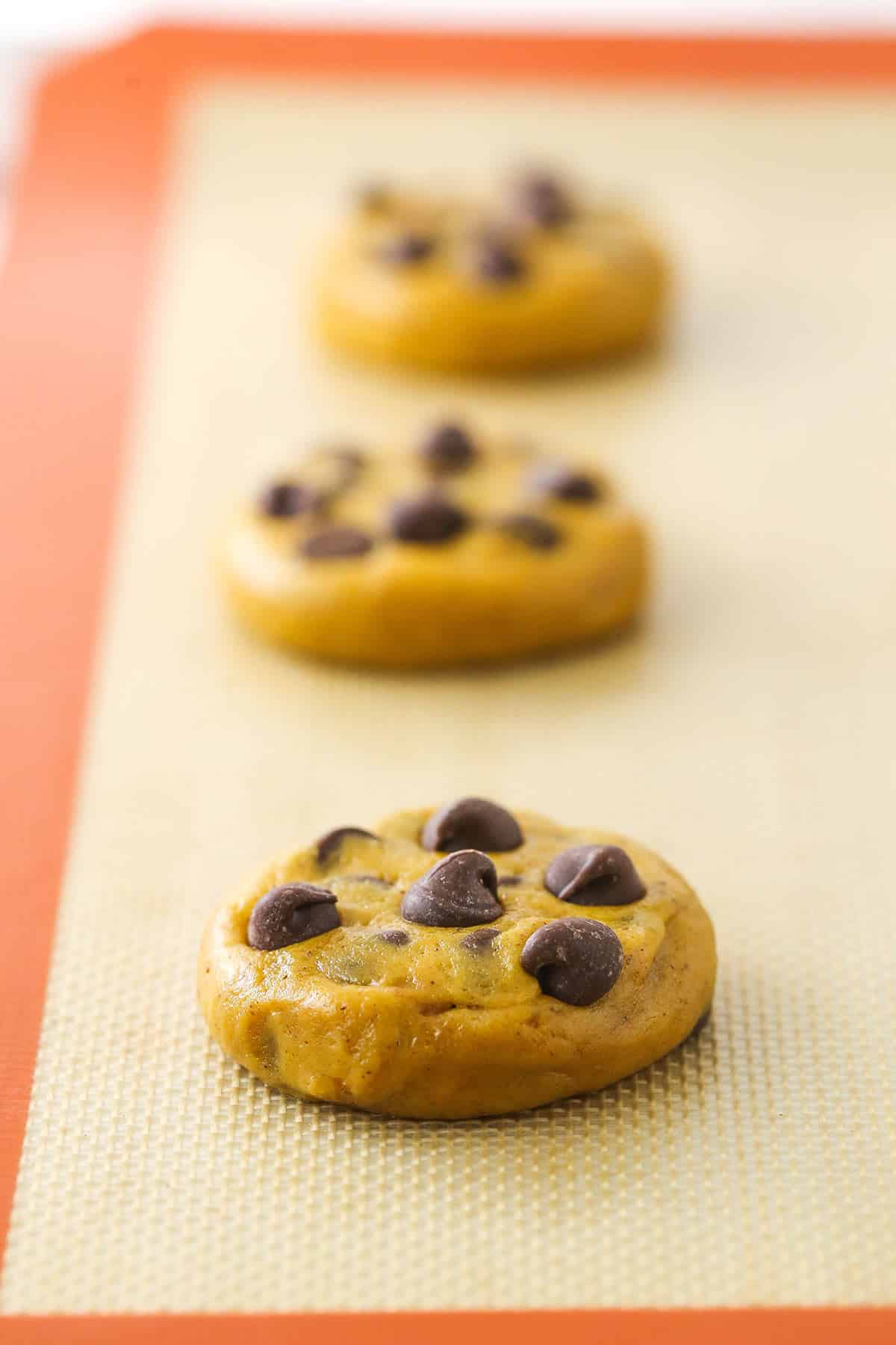 Three cookie dough balls pressed down onto a cookie sheet lined with a baking mat.