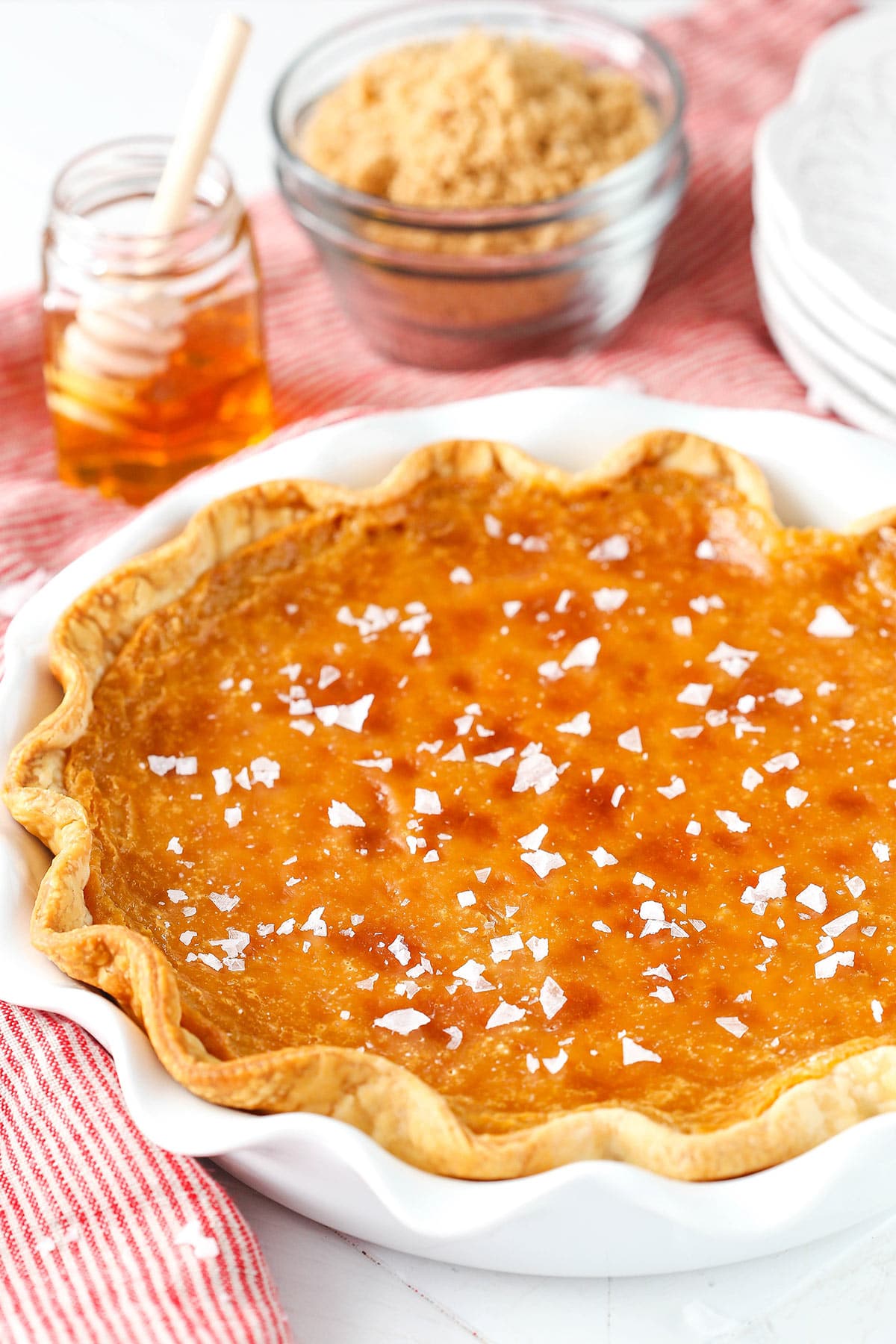 A salted honey pie on a table beside a striped towel, a jar of honey and a bowl of light brown sugar