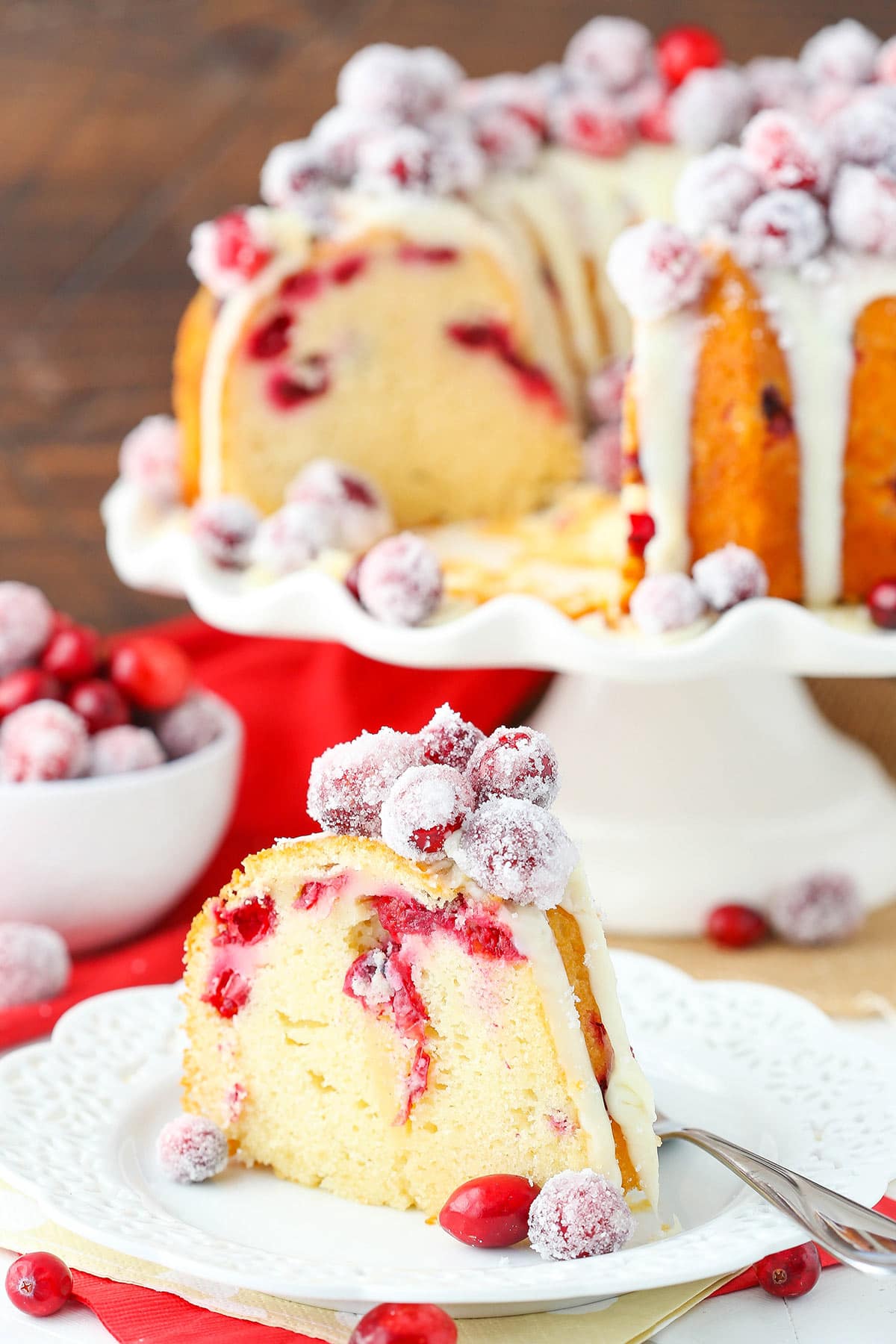 A slice of sparkling cranberry bundt cake on a plate with the remaining cake on a stand in the background