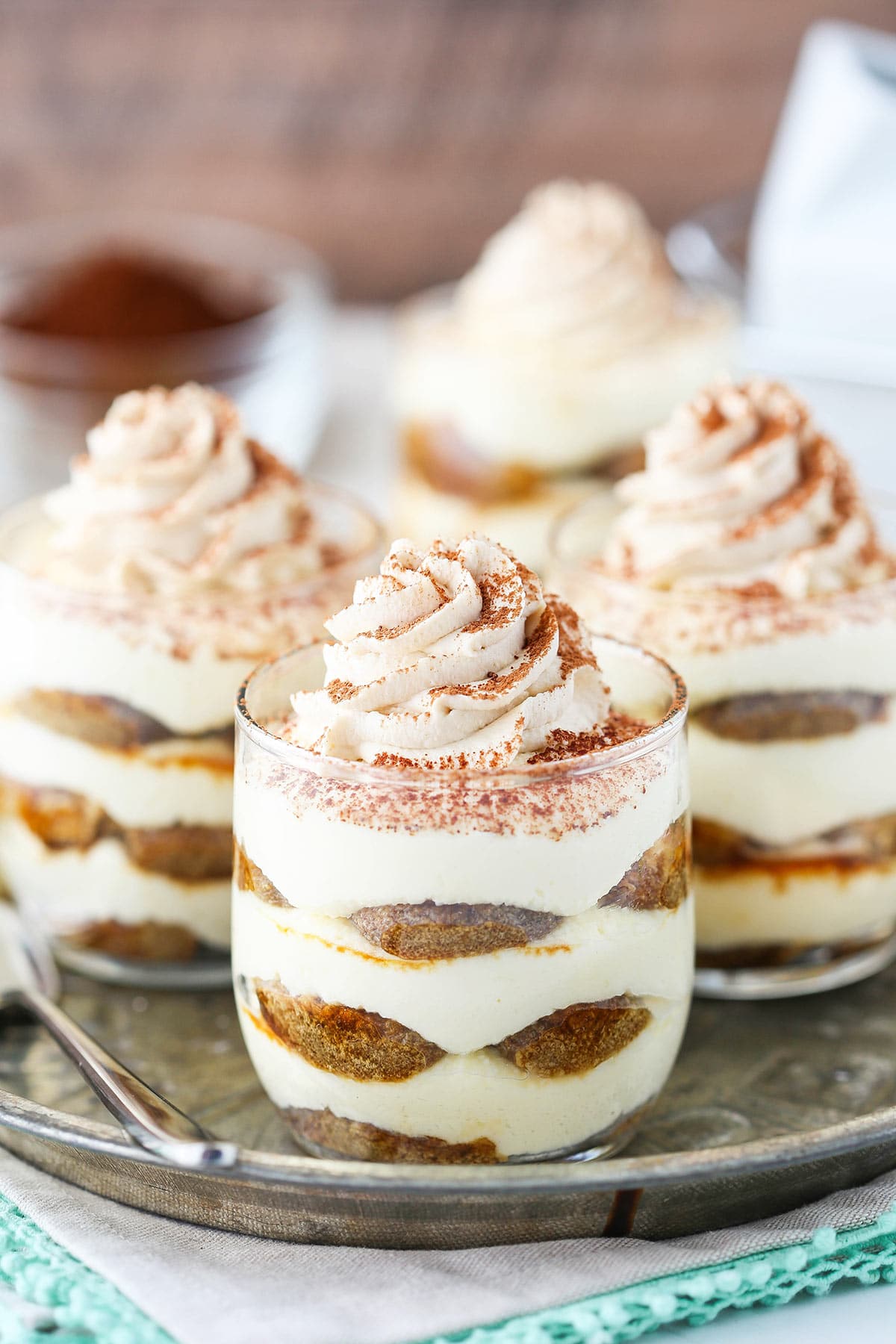 A metal tray holding three tiramisu trifles with another trifle and a dish full of cocoa powder behind the tray