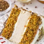 slice of carrot cake cheesecake cake on white plate with pecans, pineapple and carrots in bowls in the background