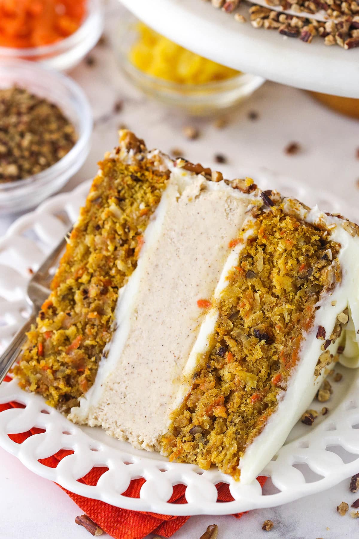This Carrot Cake Cheesecake Cake has two layers of super moist carrot cake and a layer of creamy cinnamon cheesecake! Covered in a homemade cream cheese frosting, it's the perfect cake for Easter! slice of carrot cake cheesecake cake on white plate with pecans, pineapple and carrots in bowls in the background