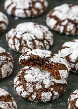 Assorted chocolate crinkle cookies on a dark grey surface, with one cookie with a bite missing stacked on top of another cookie.