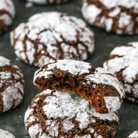 Assorted chocolate crinkle cookies on a dark grey surface, with one cookie with a bite missing stacked on top of another cookie.