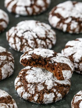 Assorted chocolate crinkle cookies on a dark grey surface, with one cookie with a bite missing stacked on top of another cookie.