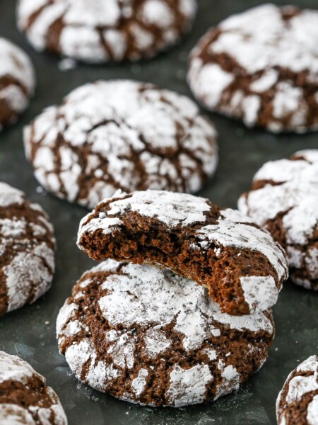 Assorted chocolate crinkle cookies on a dark grey surface, with one cookie with a bite missing stacked on top of another cookie.