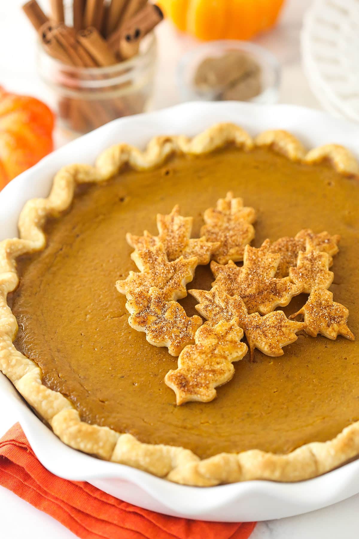 Pumpkin pie with leaf pie toppers on a orange napkin with cinnamon sticks and pumpkins in the background