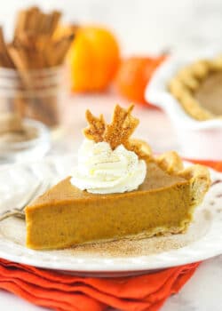 a slice of pumpkin pie on a white plate on an orange napkin with whipped cream and pie crust leaf on top