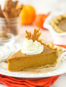 a slice of pumpkin pie on a white plate on an orange napkin with whipped cream and pie crust leaf on top