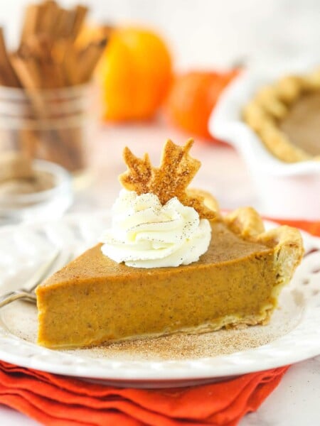 a slice of pumpkin pie on a white plate on an orange napkin with whipped cream and pie crust leaf on top