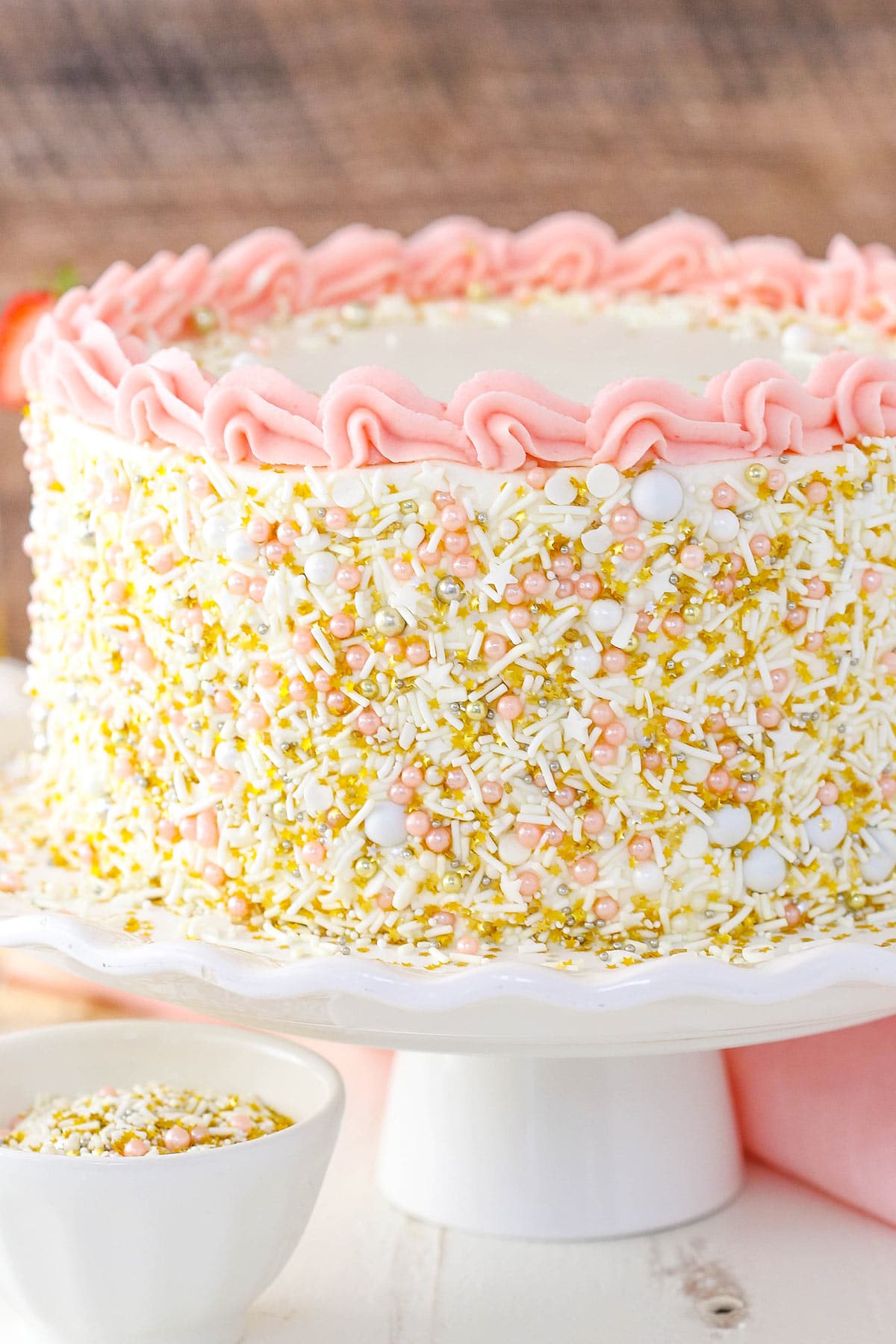 A strawberry champagne layer cake on a white cake stand beside a pink kitchen towel