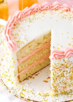 A champagne layer cake on a cardboard cake round with one large slice removed