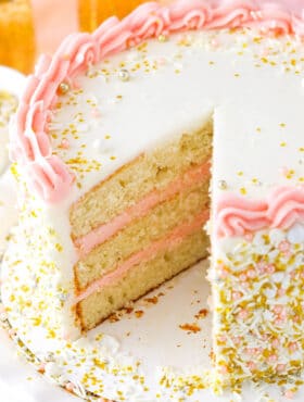 A champagne layer cake on a cardboard cake round with one large slice removed