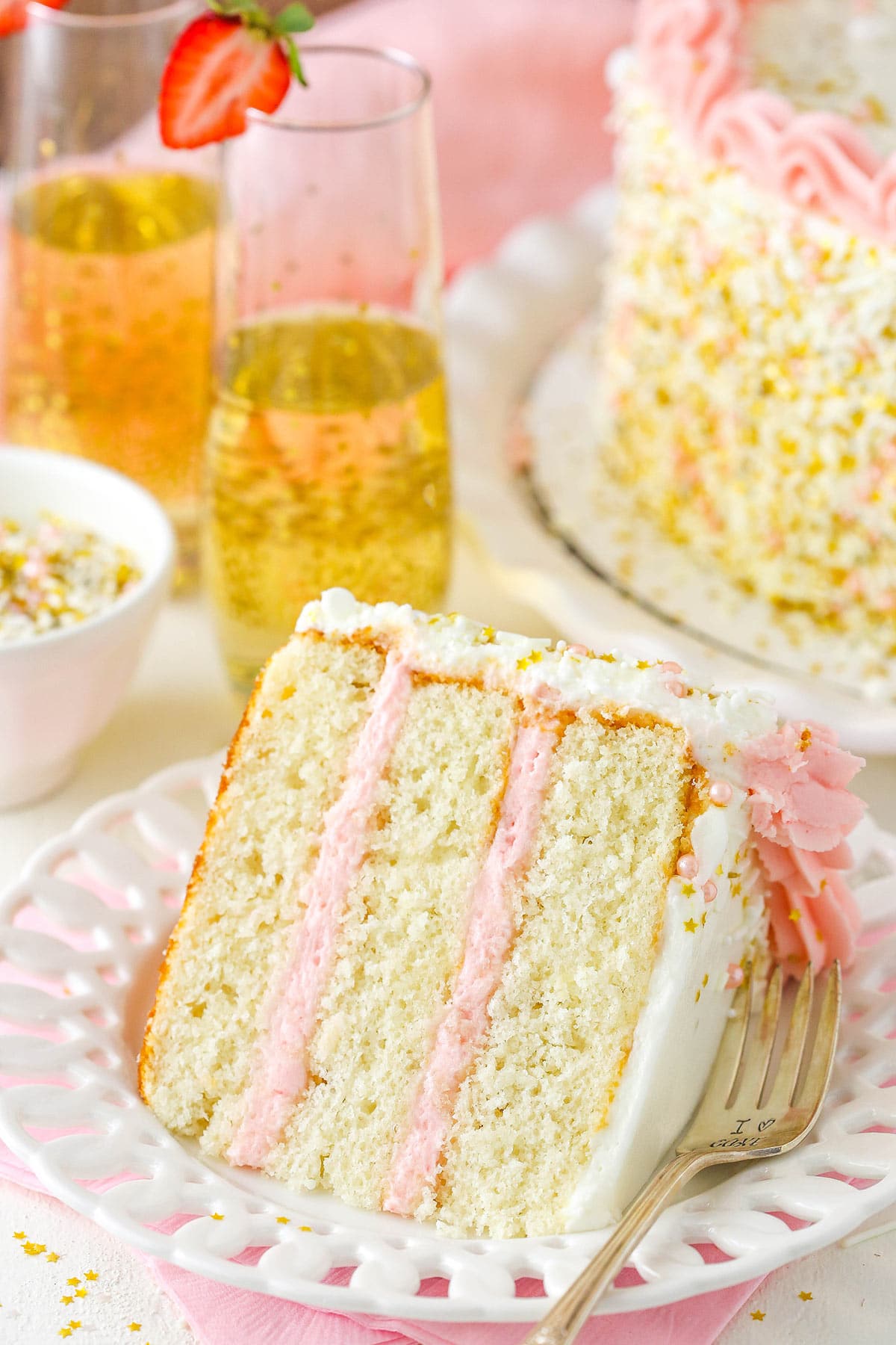 A slice of strawberry champagne cake on a plate with two glasses of champagne behind it