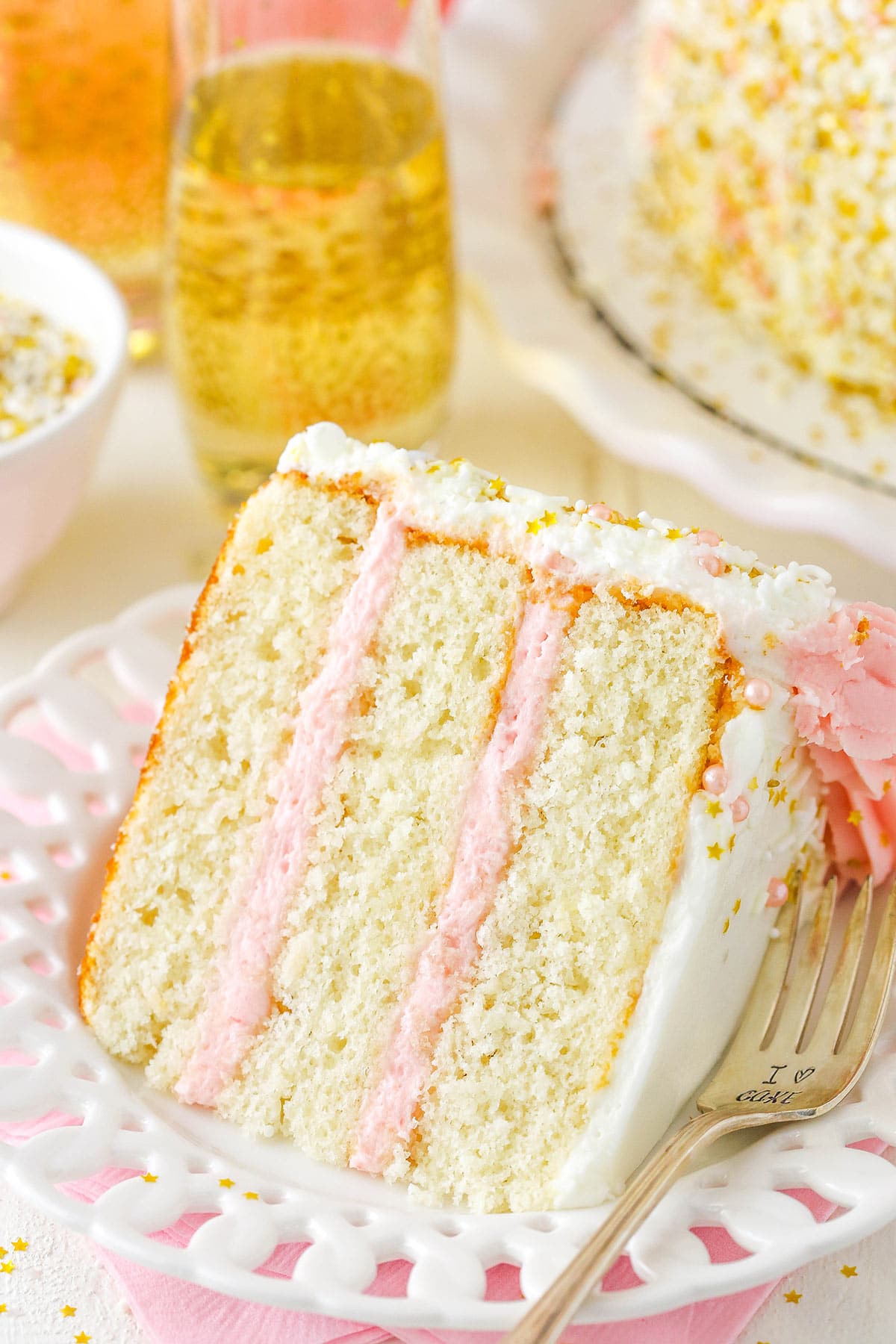 A slice of strawberry champagne layer cake on a plate with the remaining cake visible in the background