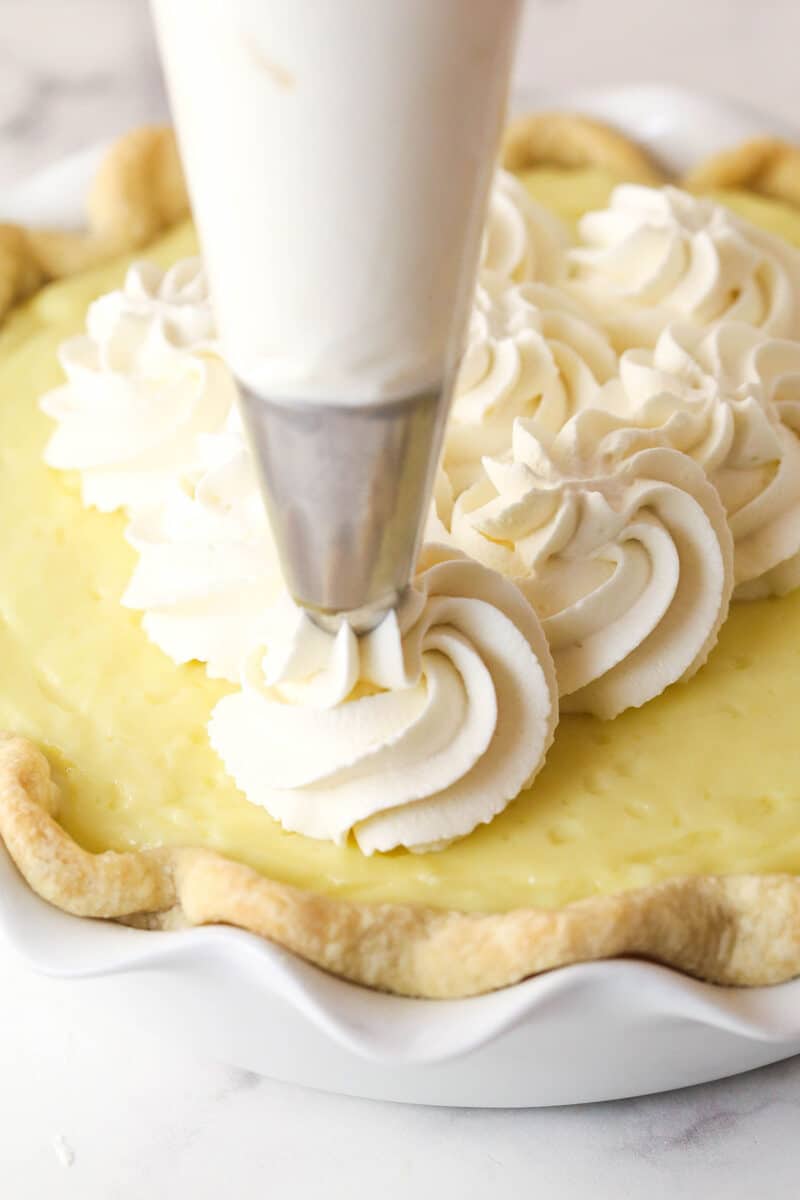 This coconut cream pie is a classic, creamy dessert made from rich coconut custard filling topped with whipped cream in a buttery pie crust. Piping whipped cream onto coconut cream pie.