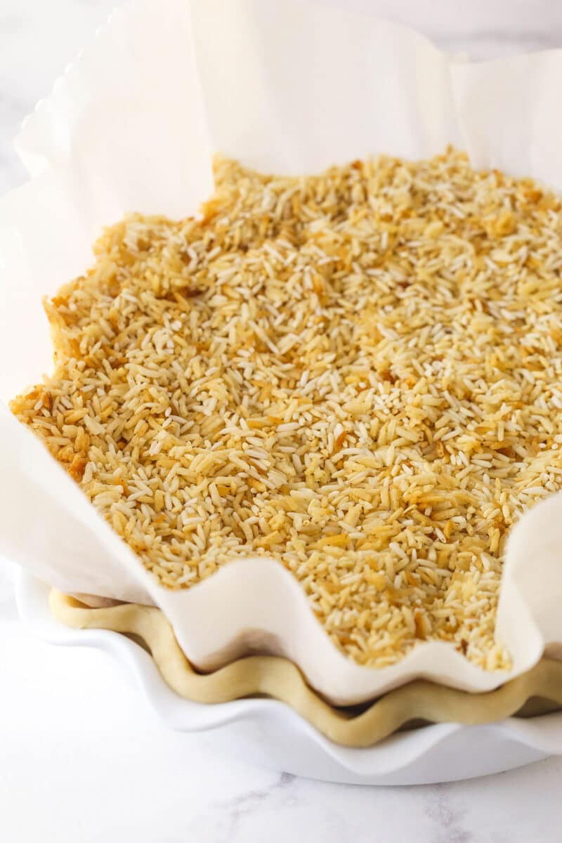 This coconut cream pie is a classic, creamy dessert made from rich coconut custard filling topped with whipped cream in a buttery pie crust. Rice weighing down parchment paper inside a par-baked pie crust.