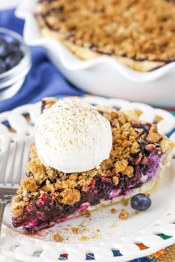 A slice of Blueberry Crumb Cheesecake Pie on a white plate with a scoop of ice cream on top. 