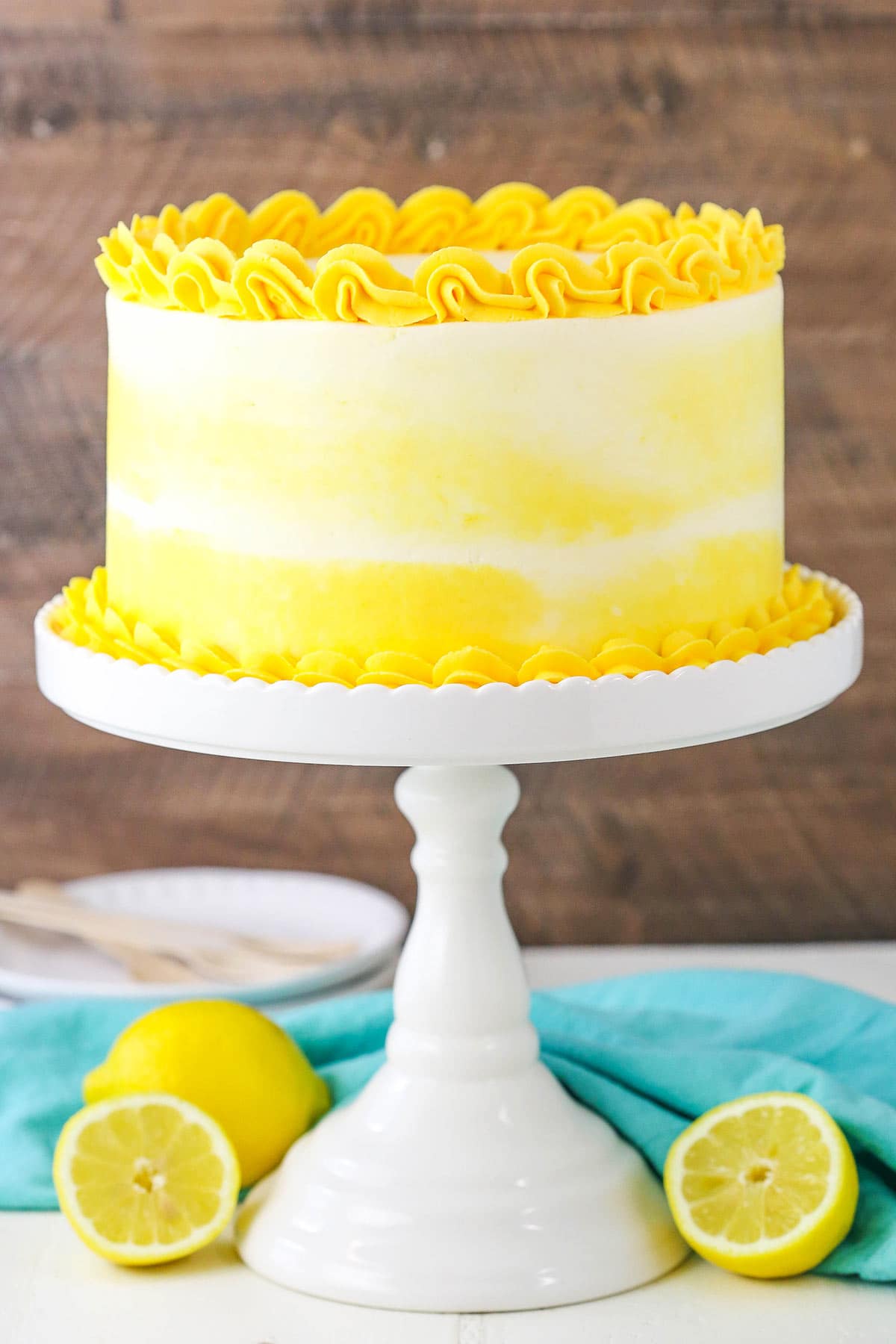 A Lemon Layer Cake on a Tall White Cake Stand Sitting on a Table with Fresh Lemons