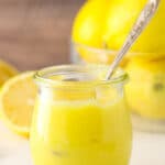 Lemon curd with a spoon in a jar near a bowl of lemons and a halved lemon.