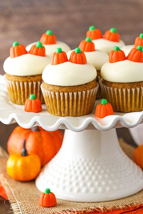 Seven cupcakes with cream cheese frosting on a cake stand beside two pumpkins