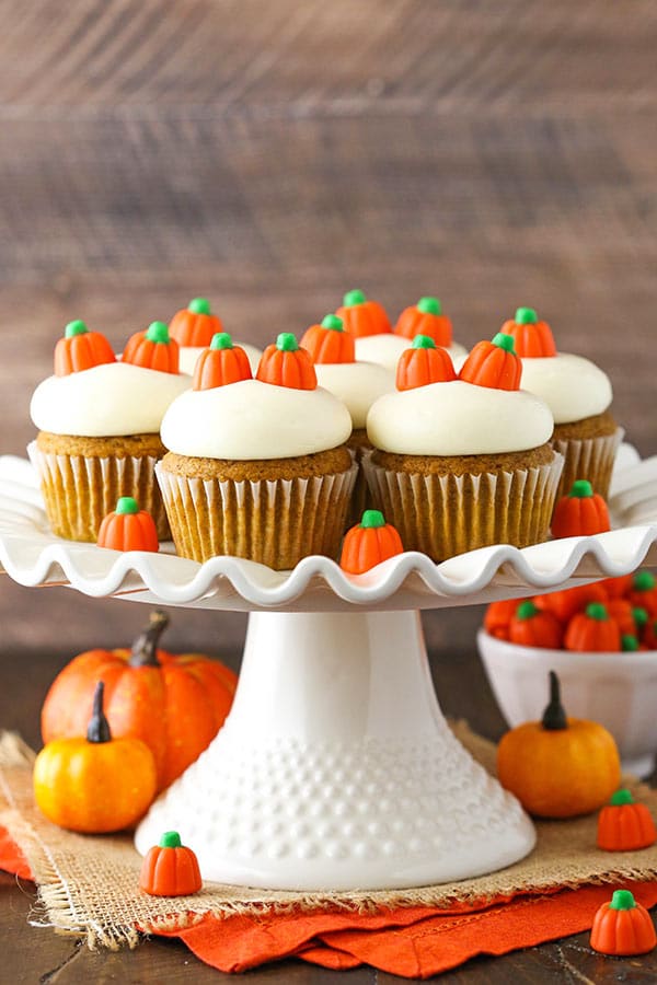 A white cake stand on a table holding seven homemade pumpkin cupcakes