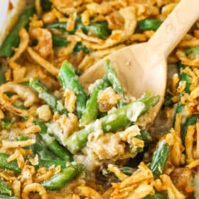 A wooden spoon scooping up a serving of green bean casserole from a ceramic baking dish.