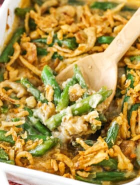 A wooden spoon scooping up a serving of green bean casserole from a ceramic baking dish.