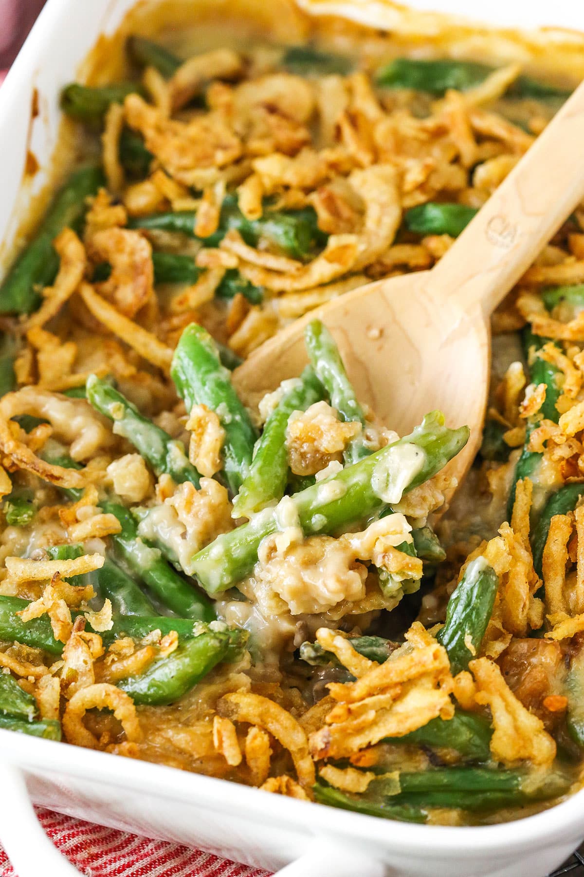 With creamy cheese, crispy fried onions, and Campbell's cream of mushroom, this classic green bean casserole recipe hits all the right notes! A wooden spoon scooping up a serving of green bean casserole from a ceramic baking dish.