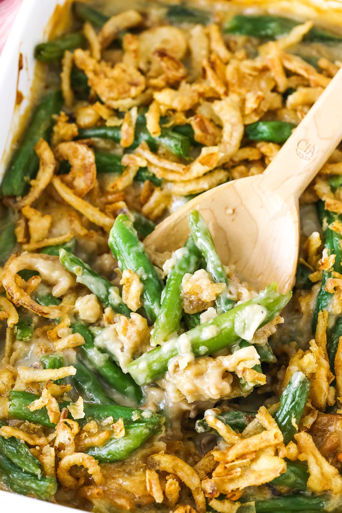 A wooden spoon scooping up a serving of green bean casserole from a ceramic baking dish.