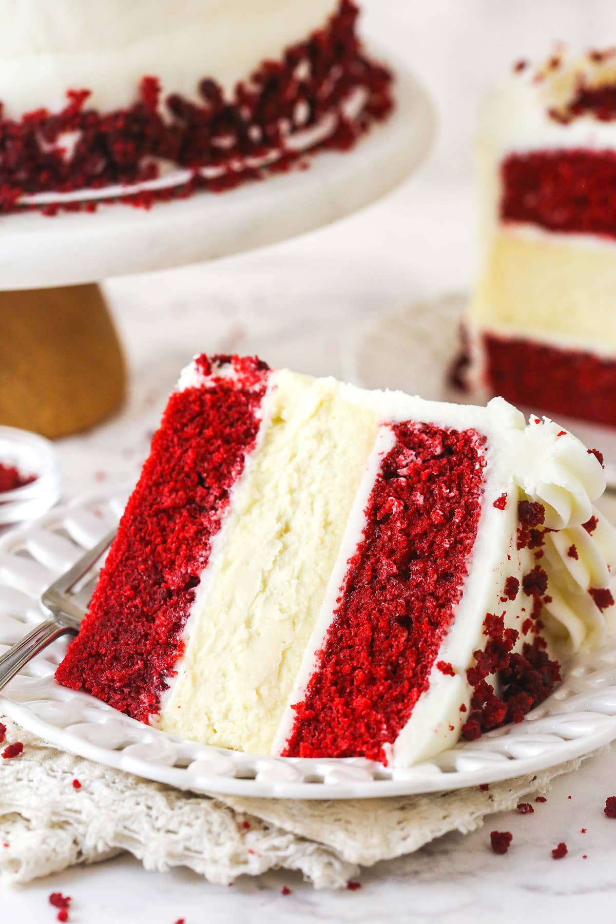 piece of red velvet cheesecake cake on white plate, cream napkin and marble table