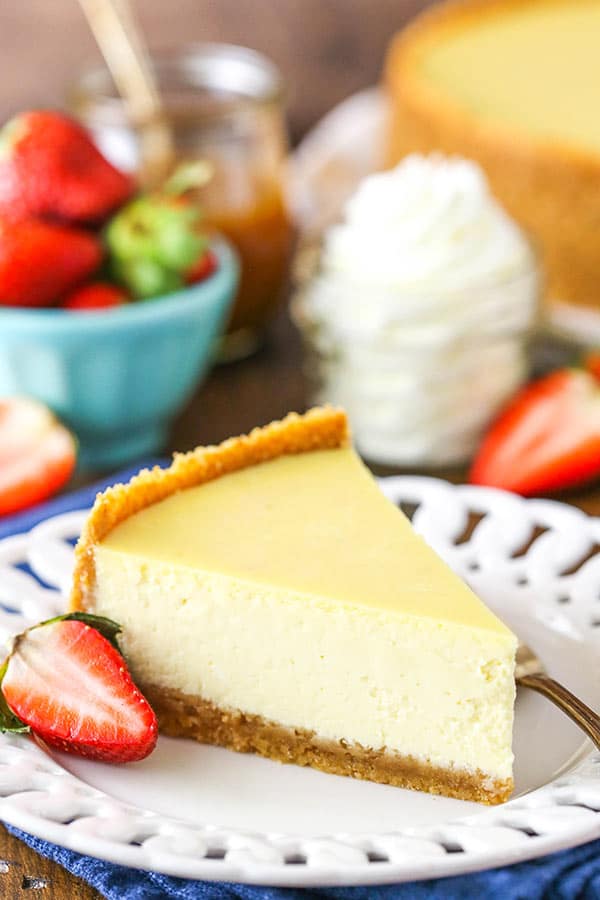 A slice of creamy cheesecake on a white plate next to a sliced strawberry