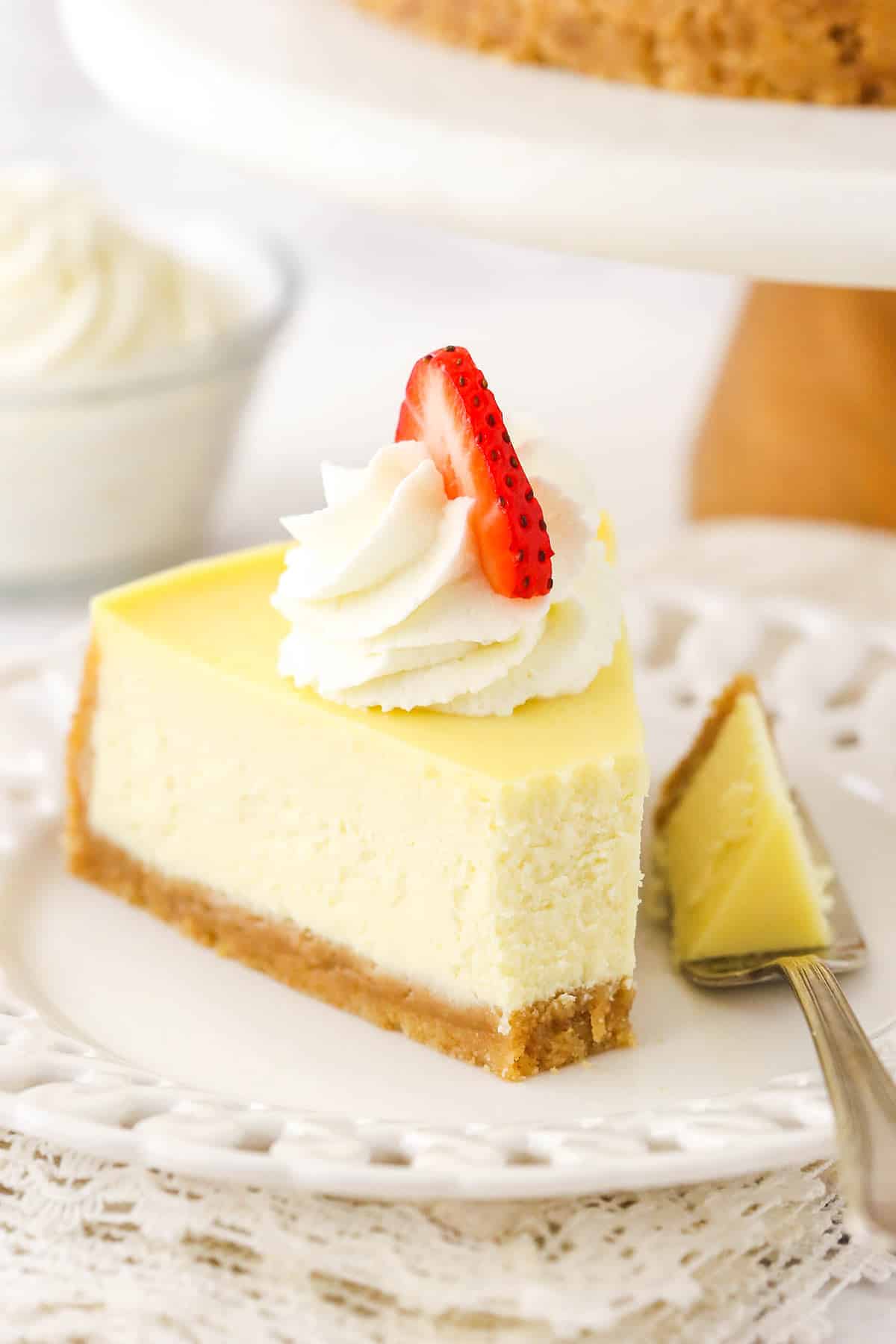 A slice of homemade cheese cake with whipped cream and strawberries, a bite taken out of the cheesecake with a fork next to it