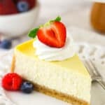 A slice of homemade cheesecake with whipped cream and berries with a bowl of berries in the background