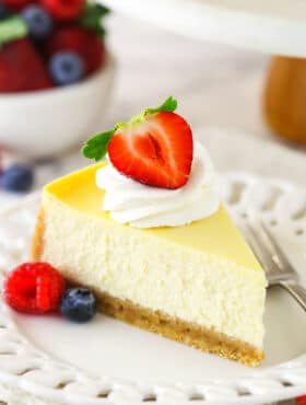 A slice of homemade cheesecake with whipped cream and berries with a bowl of berries in the background