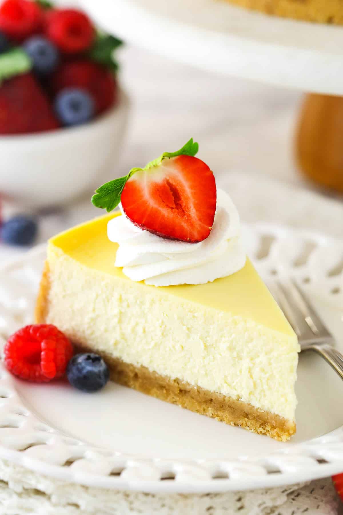 A slice of homemade cheesecake with whipped cream and berries with a bowl of berries in the background