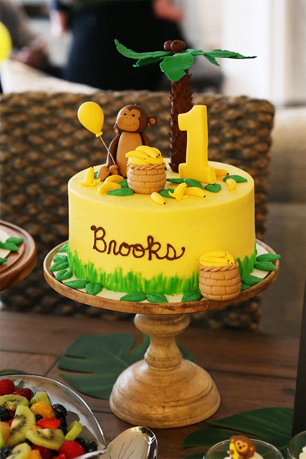 Brooks' First Birthday Cake on a Light Wooden Cake Stand Beside a Bowl of Fruit Salad