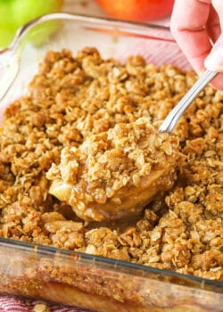 An apple crisp in a glass baking dish with a serving spoon removing a heaping section from the middle