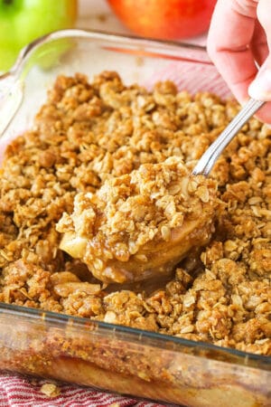 An apple crisp in a glass baking dish with a serving spoon removing a heaping section from the middle
