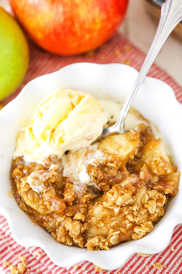 A bowl of apple crisp with ice cream in front of two apples, one red and one green
