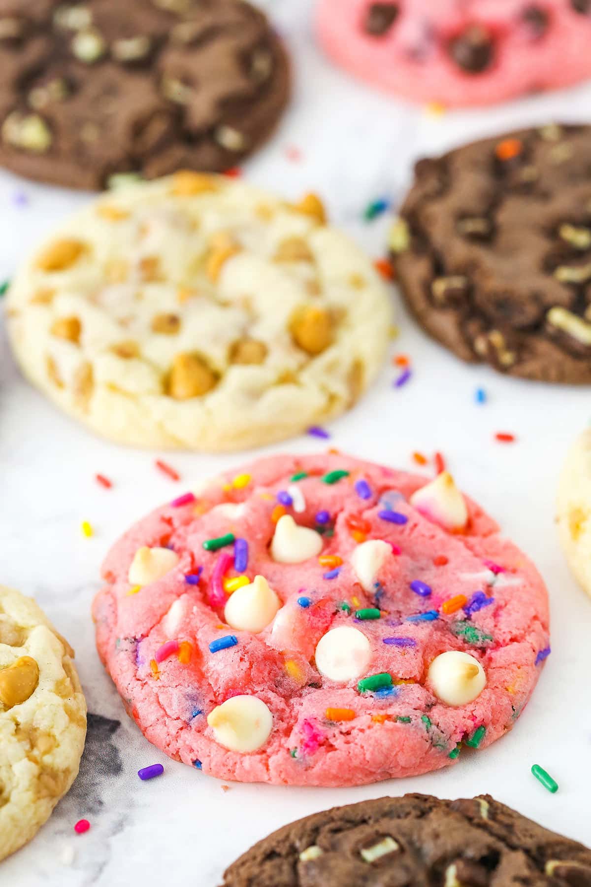 A strawberry Funfetti white chocolate chip cookie on a marble countertop beside more cookies of various flavors