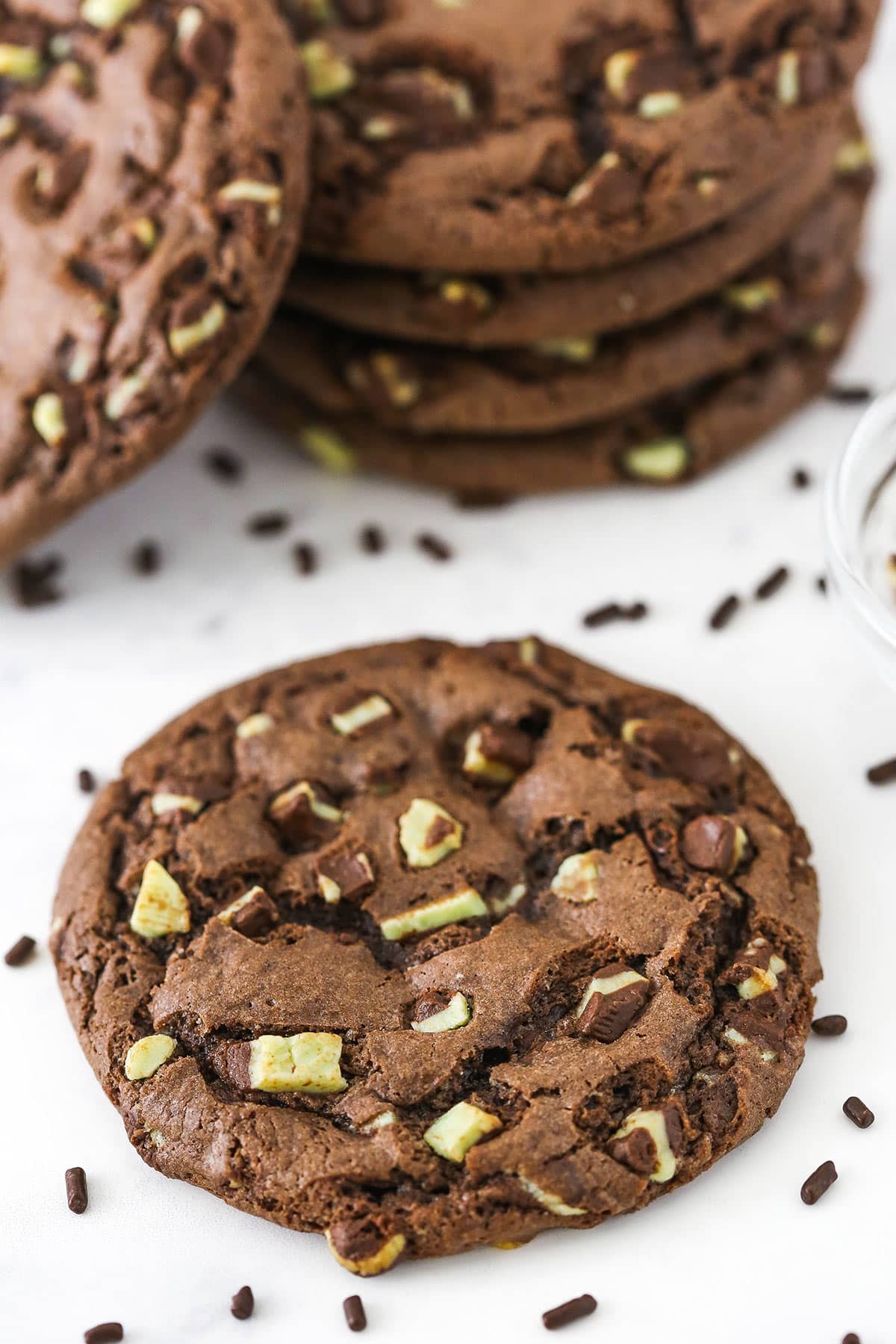 A mint chocolate chip cookie on a white surface with chocolate sprinkles scattered about