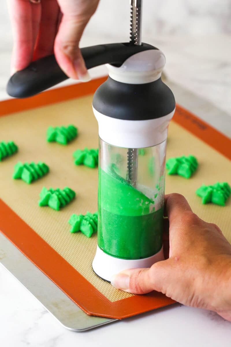 Spritz cookies are buttery, classic Christmas cookies made with a cookie press. This recipe is easy to make, with so many ways to decorate! Using a cookie press to press spritz cookies in green Christmas tree shapes onto a baking mat.