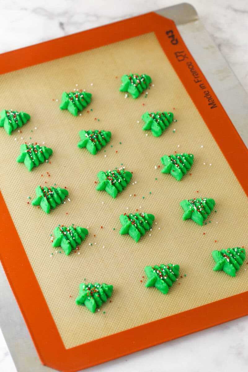 Spritz cookies are buttery, classic Christmas cookies made with a cookie press. This recipe is easy to make, with so many ways to decorate! Spritz cookies pressed out into green Christmas tree shapes on a silicone baking mat.