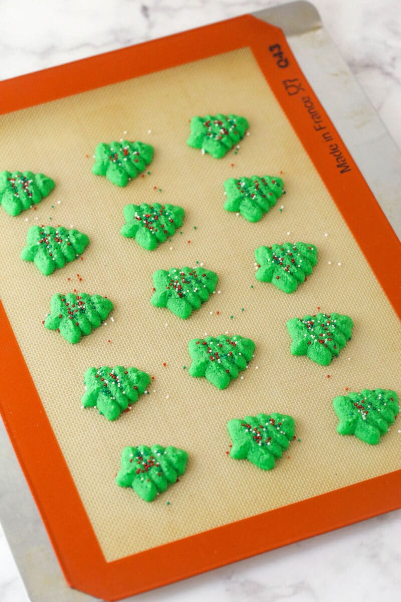 Spritz cookies are buttery, classic Christmas cookies made with a cookie press. This recipe is easy to make, with so many ways to decorate! Baked Christmas tree spritz cookies on a silicone baking mat.