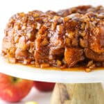 Full Apple Fritter Monkey Bread on a white cake stand with chopped apples in the background