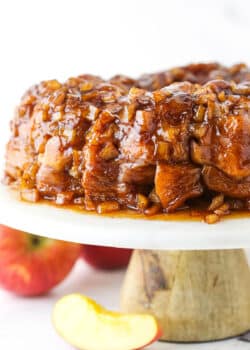 Full Apple Fritter Monkey Bread on a white cake stand with chopped apples in the background