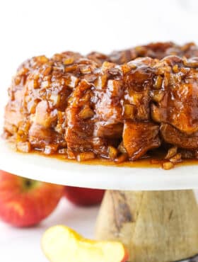 Full Apple Fritter Monkey Bread on a white cake stand with chopped apples in the background