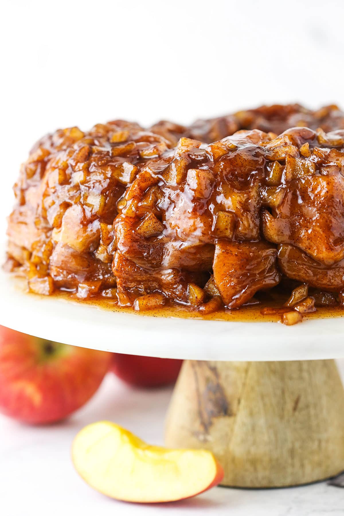 Full Apple Fritter Monkey Bread on a white cake stand with chopped apples in the background